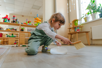 nursery child using a toy dustpan and brush to sweep floor of playroom